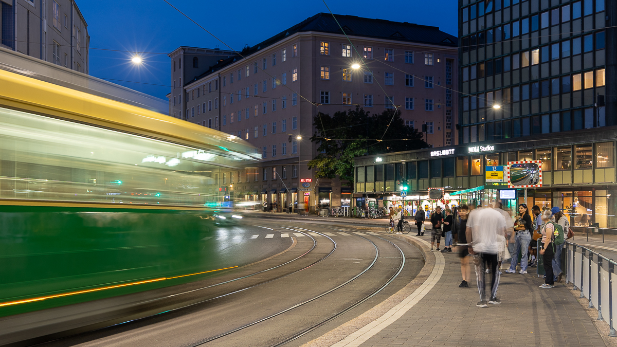 Ihmisiä iltamyöhään raitiovaunupysäkillä, raitiovaunu näkyy liikkeen vuoksi värijuovana.